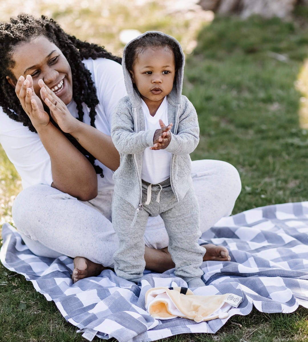 A child wearing a gray hoodie and sweatpants stands on a picnic blanket with a woman nearby clapping her hands.