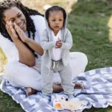 A child wearing a gray hoodie and sweatpants stands on a picnic blanket with a woman nearby clapping her hands.