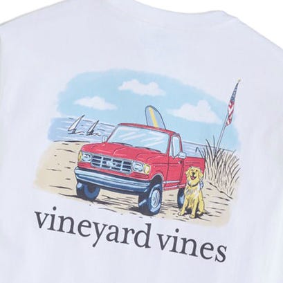 A white Vineyard Vines shirt features an illustration of a red pickup truck with a surfboard, a dog, and an American flag on a beach with sailboats in the background.