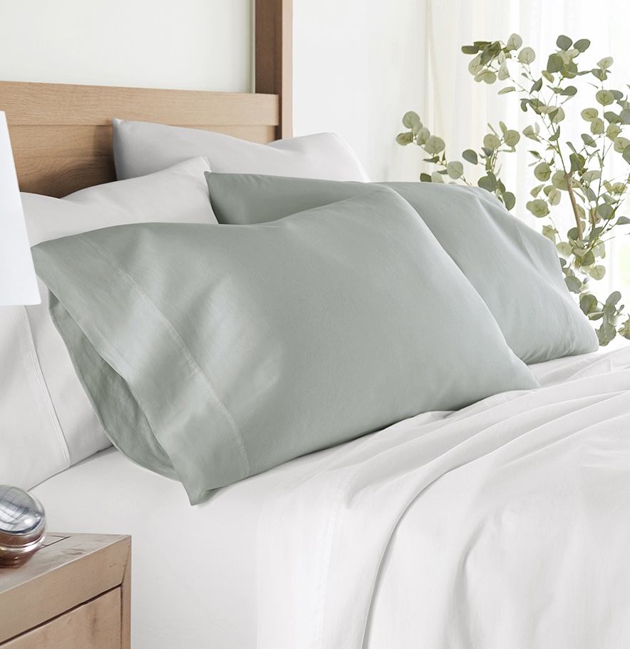 A neatly made bed with sage green pillowcases and white sheets, alongside a wooden nightstand and a potted eucalyptus plant.