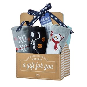 A 6-piece holiday hand towel gift set is displayed in a cardboard box labeled \“Cheers! a gift for you.\“ The set includes towels with festive patterns like arrows, a skeleton, and a snowman. A blue tag labeled \“Holidays Hand Towels\“ is attached with a ribbon.