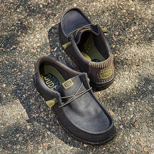 A pair of casual black slip-on shoes with green accents and laces, featuring \“Hey Dude\“ branding on the insole and heel, displayed on a textured outdoor surface.