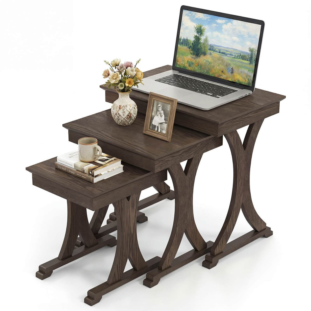 A set of three dark wood nesting tables with curved legs, each differing in height. The largest table holds a laptop, a vase of flowers, and a framed photo, while the smallest has a coffee mug and stacked books.
