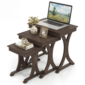 A set of three dark wood nesting tables with curved legs, each differing in height. The largest table holds a laptop, a vase of flowers, and a framed photo, while the smallest has a coffee mug and stacked books.