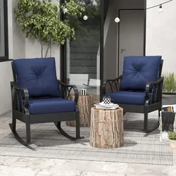Two blue cushioned rocking chairs with black frames sit on a patterned outdoor rug, accompanied by round wooden stump-style side tables, outdoor string lights, and potted plants on a terrace.