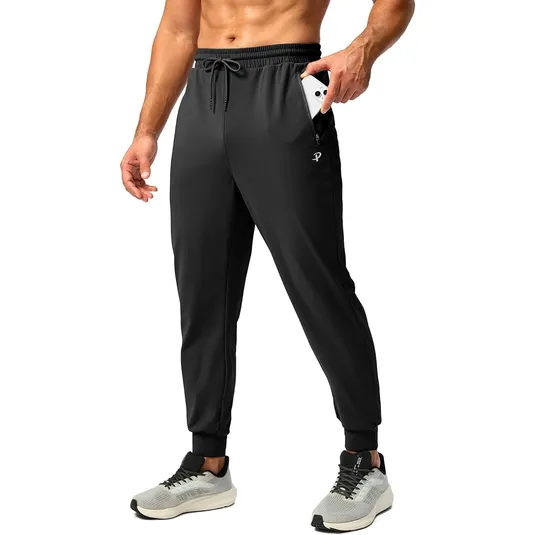 Black athletic joggers with a drawstring waist and zippered side pocket, shown holding a smartphone. Paired with light grey and black athletic shoes.
