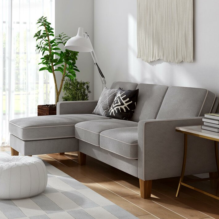 A compact, gray sectional sofa with a reversible chaise, paired with a geometric throw pillow and wooden legs, set on a light hardwood floor. It features a white floor lamp, wall hanging, and greenery nearby, creating a cozy and modern living space.