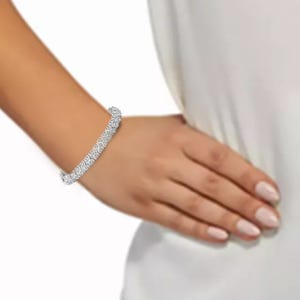 A wrist adorned with a silver bracelet featuring a series of intricate, sparkling gemstones in a linked pattern. The arm rests on a white fabric background.