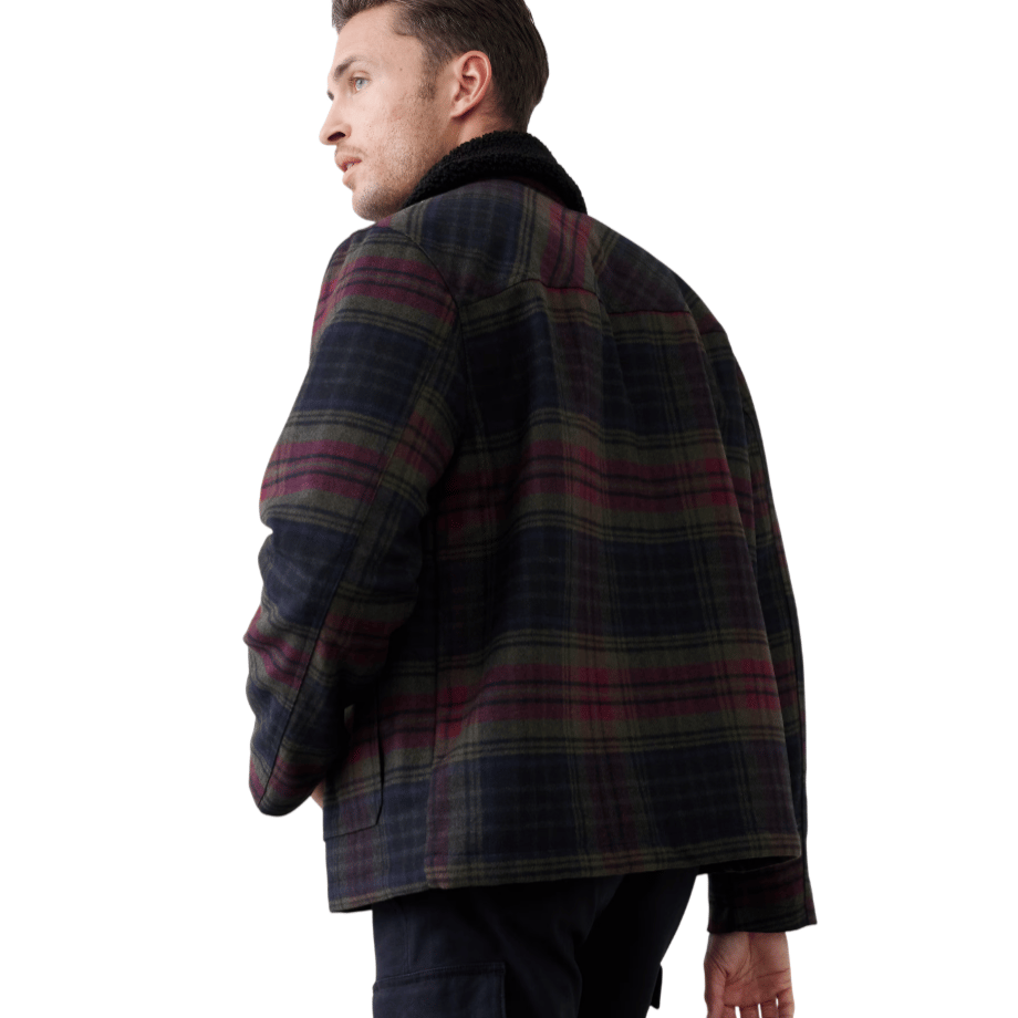 A man is wearing a plaid jacket with dark maroon and green tones, featuring a black shearling collar, paired with dark cargo pants.