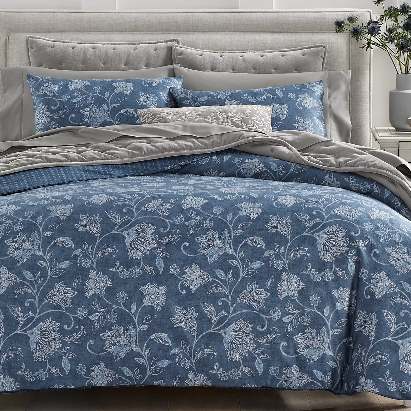 A bedding set featuring a blue floral pattern includes pillows and a comforter on a tufted headboard bed, complemented by grey accents and a decorative cushion.