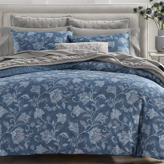 A bedding set featuring a blue floral pattern includes pillows and a comforter on a tufted headboard bed, complemented by grey accents and a decorative cushion.