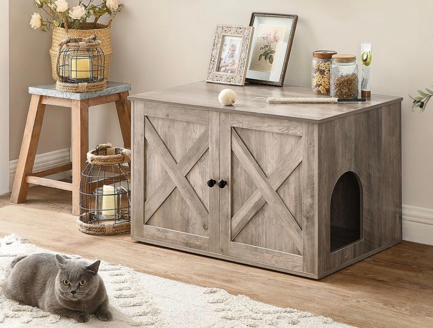 A wooden cabinet with dual doors serves as a cat litter box cover, featuring an arched entry on the side. It is accessorized with decorative items and a resting cat nearby.