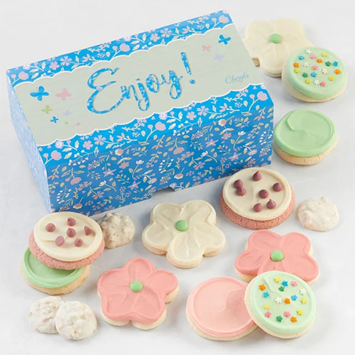 A decorative box labeled \“Enjoy!\“ contains an assortment of frosted cookies, including round and flower-shaped varieties in pastel colors with sprinkles and chocolate chips.