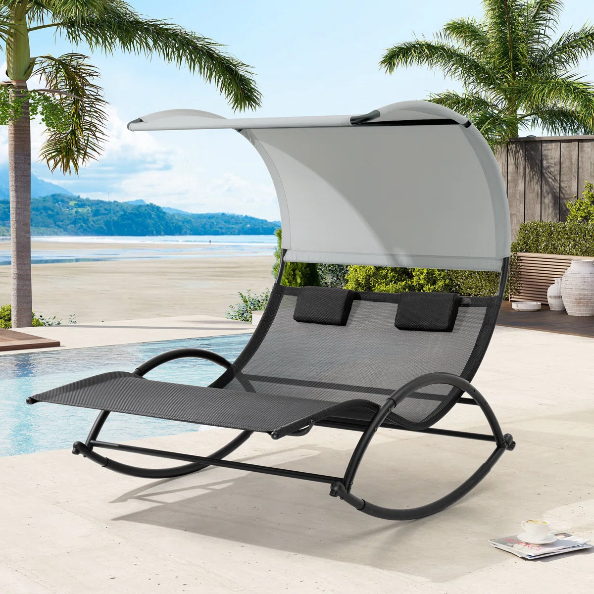 A two-person outdoor rocking chaise lounge with a sunshade canopy beside a pool, surrounded by palm trees and a beach view.
