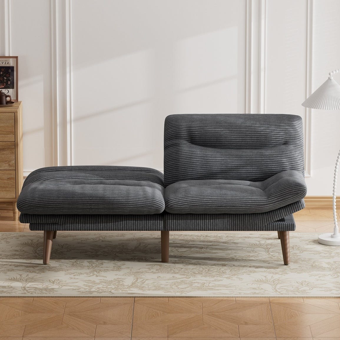 Gray corduroy chaise lounge with wooden legs set on a patterned area rug, next to a wooden drawer and a white floor lamp.