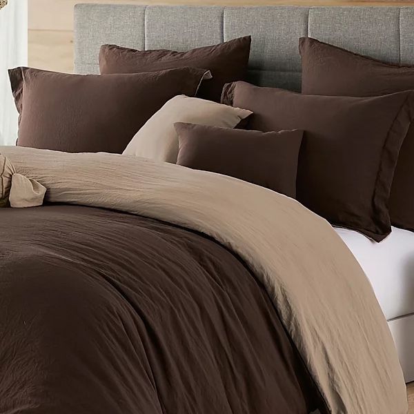 A bed with a gray upholstered headboard is dressed in brown and beige bedding, including a comforter, blankets, and multiple pillows.