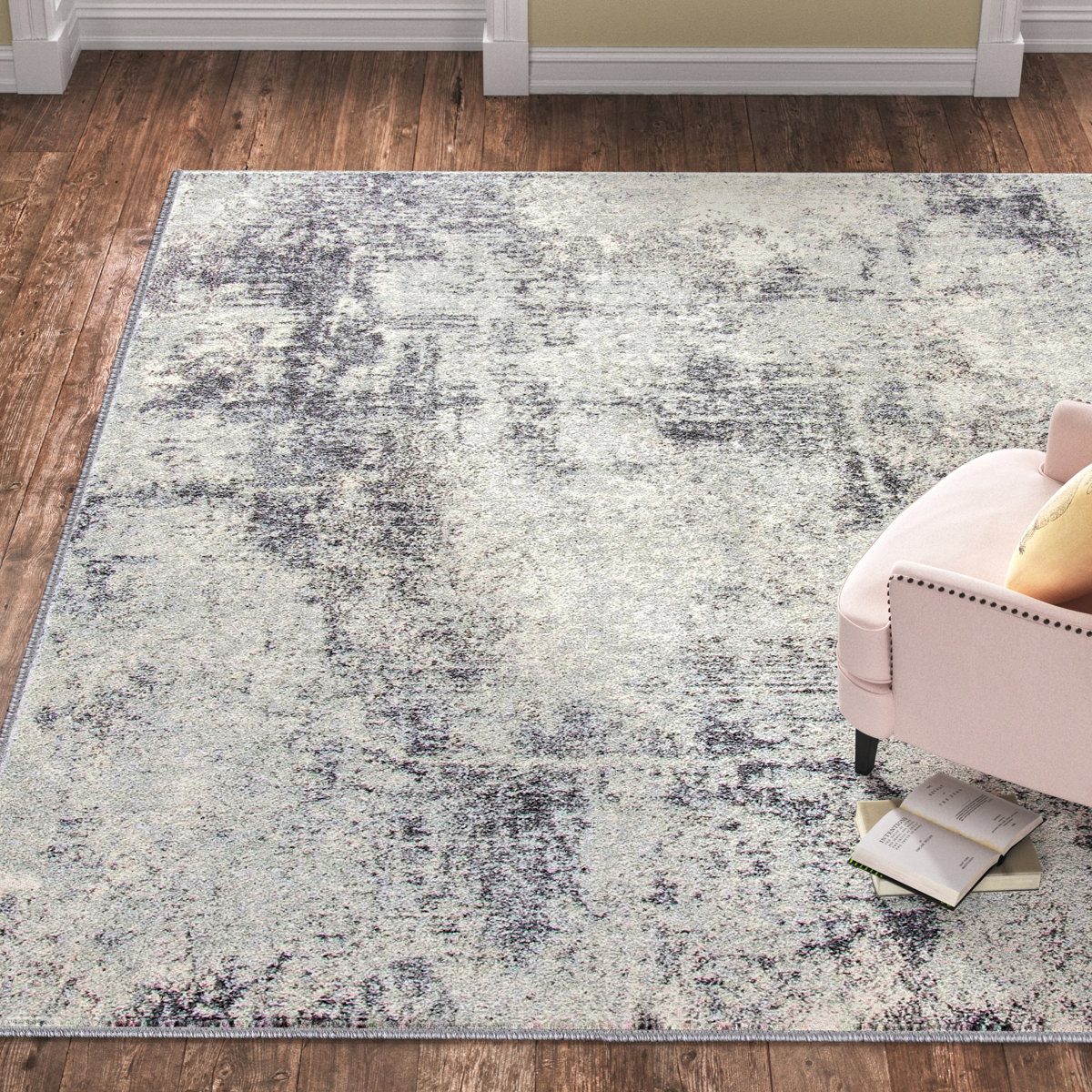 A large rectangular rug with a soft, shaggy texture and a subtle, abstract design in shades of gray and cream. It rests on a wooden floor next to a light pink chair and a few scattered books. Dimensions: 8' x 10'.
