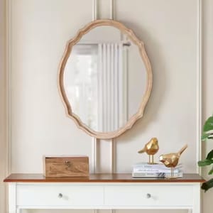 An oval mirror with a decorative, scalloped wooden frame in a natural finish, reflecting French country style. It hangs on a light-colored wall above a white console table featuring books, a wooden box, and gold bird figurines for a classic look.