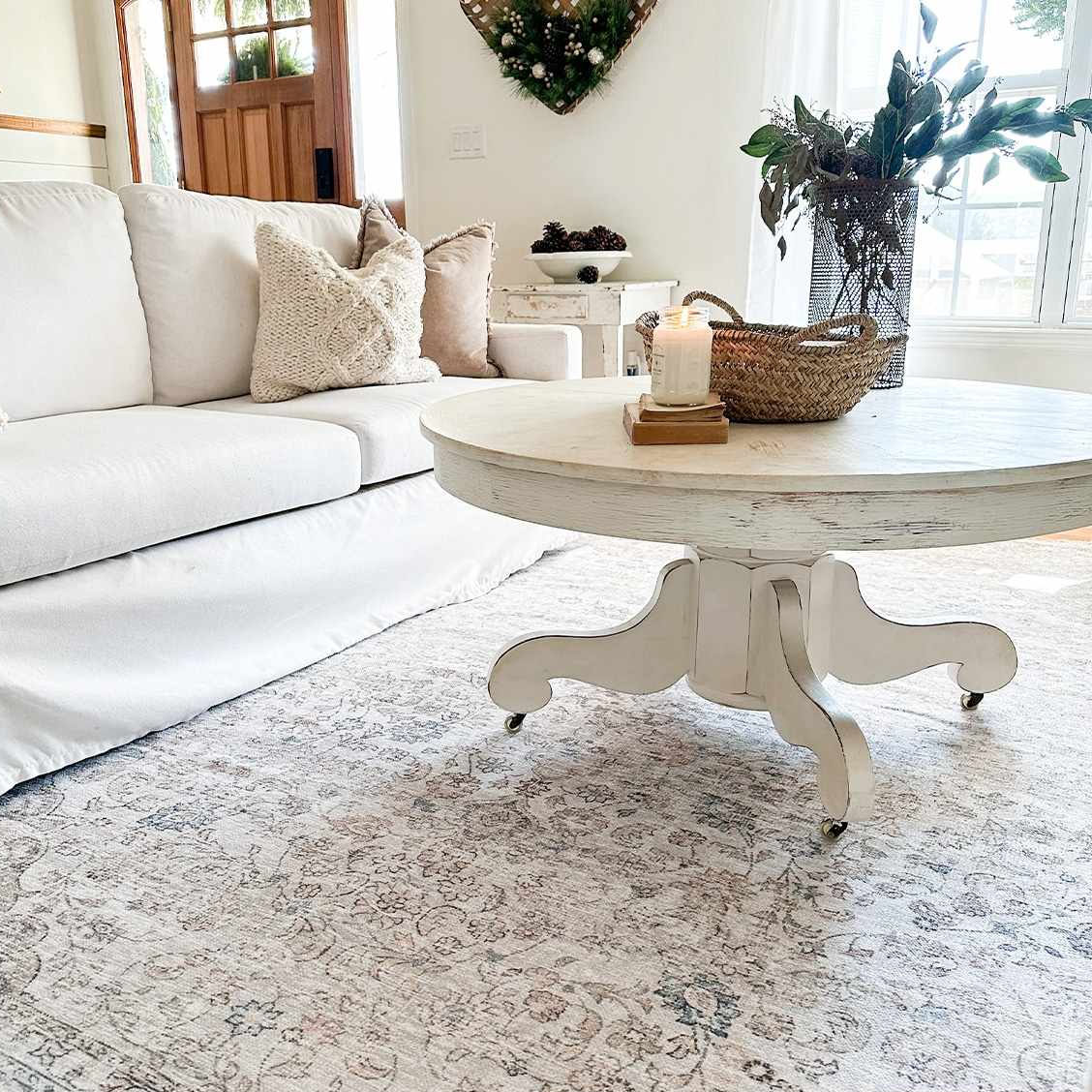 A cozy living room scene featuring a white sofa with textured pillows, a round distressed white coffee table on wheels, a woven basket with a candle, and floral decor. There's a patterned area rug and a decorative wreath on the wall.