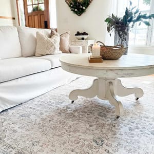 A cozy living room scene featuring a white sofa with textured pillows, a round distressed white coffee table on wheels, a woven basket with a candle, and floral decor. There's a patterned area rug and a decorative wreath on the wall.