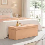Beige upholstered storage bench with a tufted lid sits at the foot of a bed in a bright bedroom, featuring large windows and neutral decor.