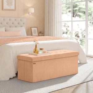 Beige upholstered storage bench with a tufted lid sits at the foot of a bed in a bright bedroom, featuring large windows and neutral decor.