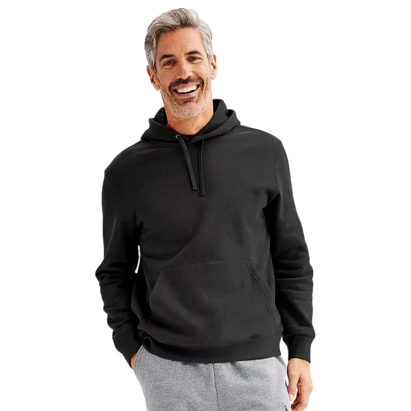 A man wearing a black pullover hoodie with a front pocket and drawstring, paired with light gray sweatpants, smiles against a plain background.