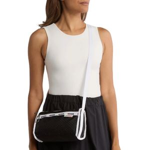 A woman carries a black crossbody bag with a quilted texture and a white strap. The bag features branded logos along the opening and is paired with a sleeveless white top and a dark skirt.