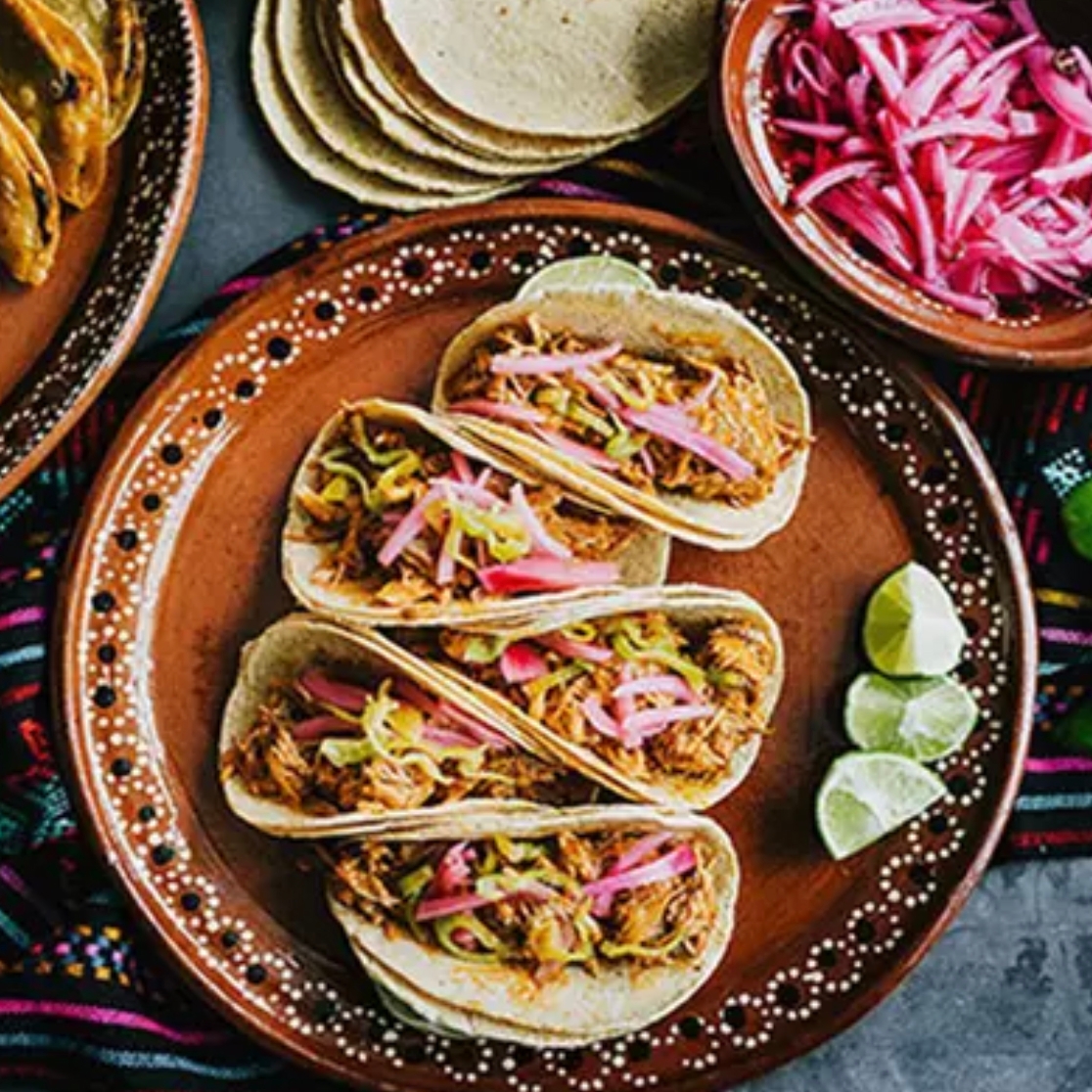 Four tacos with shredded meat and pickled onions are served on a decorative plate with lime wedges. Nearby, there's a bowl of extra pickled onions and a stack of tortillas.