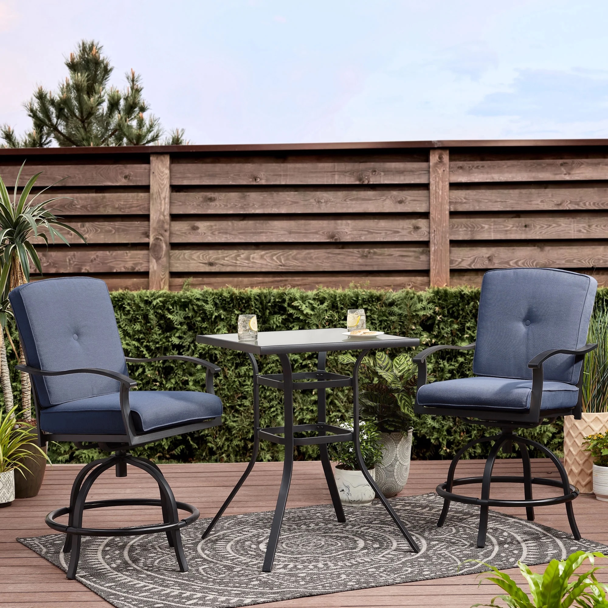 Outdoor patio set with two swivel chairs featuring blue cushions and a square table. Chairs have metal frames and footrests, placed on a deck with a patterned rug, plants, and wooden privacy fence in the background.