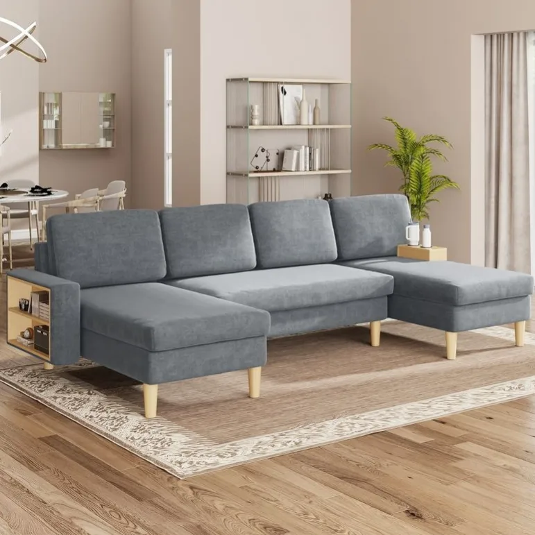 A gray sectional sofa with wooden legs, featuring built-in side storage, is placed on a decorative rug in a modern living room. The setting includes a beige wall, a bookshelf, and a potted plant.