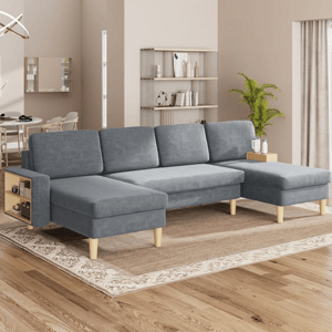 A gray sectional sofa with wooden legs, featuring built-in side storage, is placed on a decorative rug in a modern living room. The setting includes a beige wall, a bookshelf, and a potted plant.