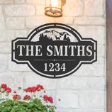 Decorative metal sign featuring mountains and trees with \“THE SMITHS\“ and \“1234,\“ mounted on a stone wall with a lantern above and a flower basket below.