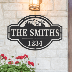 Decorative metal sign featuring mountains and trees with \“THE SMITHS\“ and \“1234,\“ mounted on a stone wall with a lantern above and a flower basket below.
