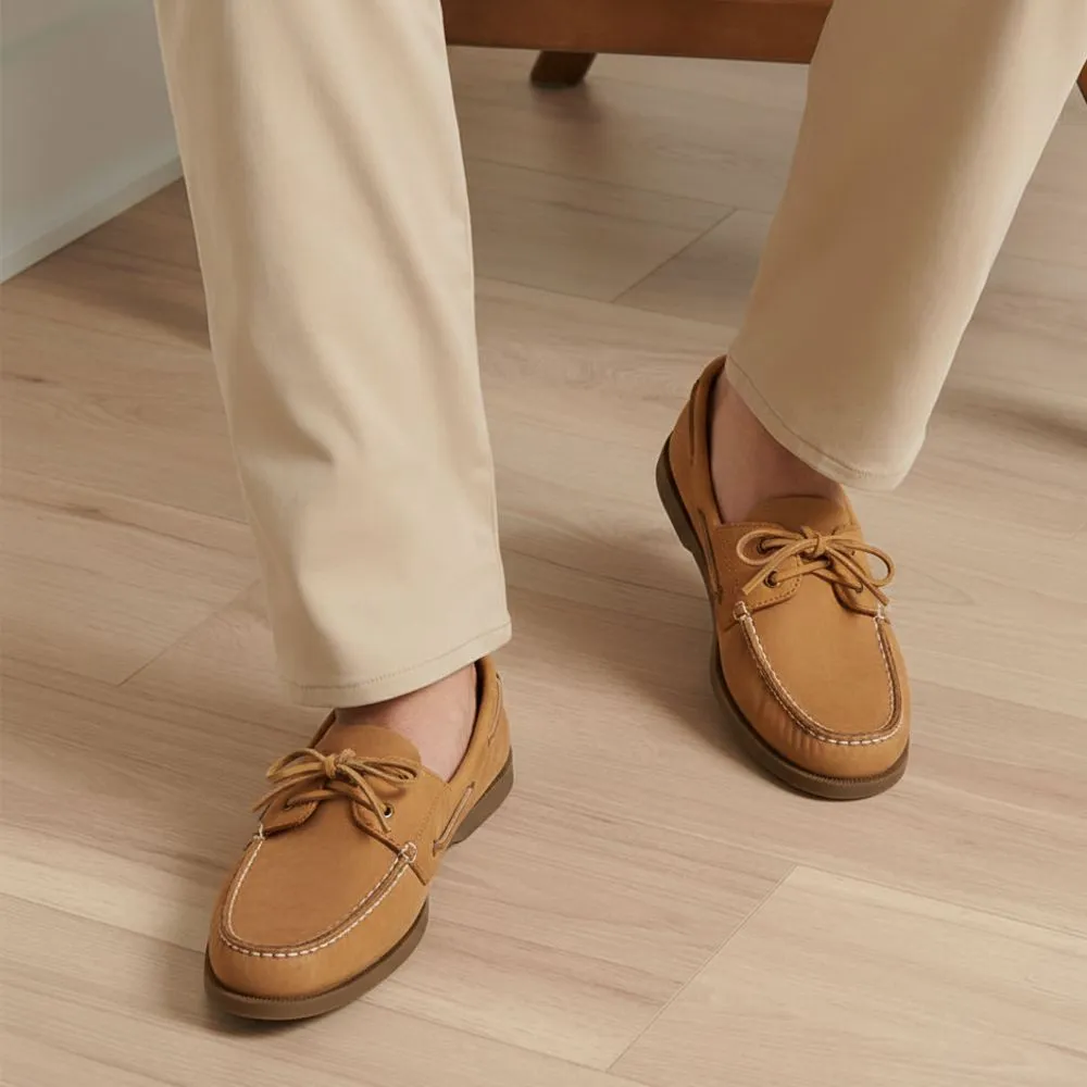 A person is wearing light brown boat shoes with dark soles and beige pants on a light wood floor. The shoes feature laces and stitching details.