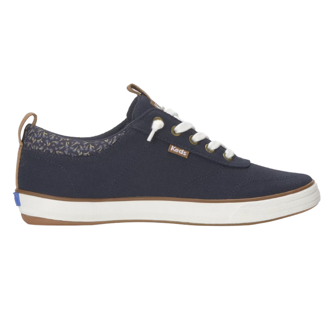 A navy blue sneaker with white laces, white sole, and brown detailing, featuring a patterned heel collar and a small \“Keds\“ logo on the side.