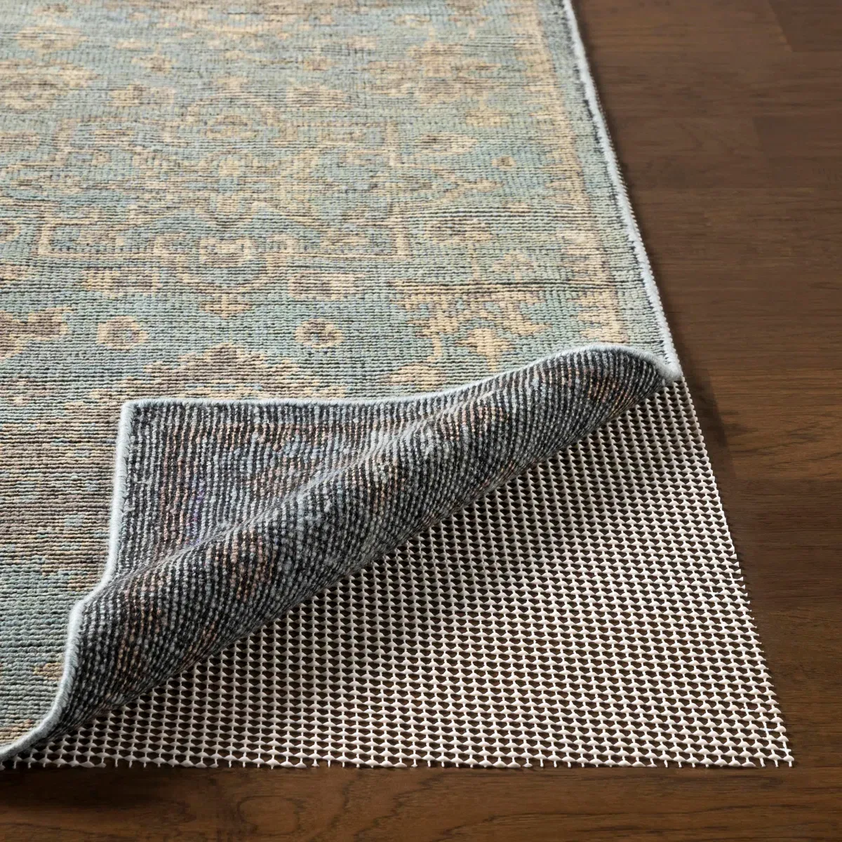 A patterned rug on a wooden floor with its corner lifted to reveal a white non-slip rug pad underneath.
