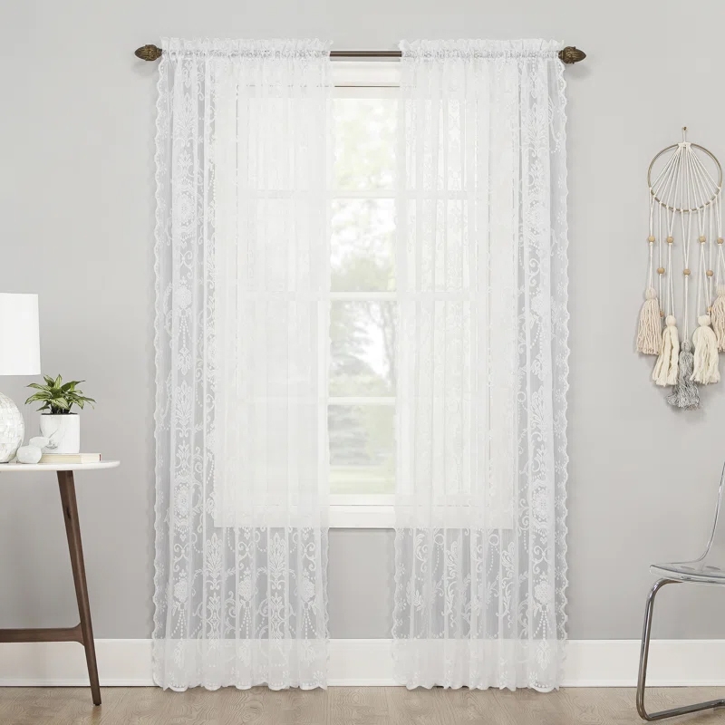 White lace curtains with a delicate, floral pattern hang on a rod over a window, flanked by a modern side table and a decorative wall hanging.