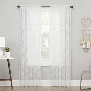 White lace curtains with a delicate, floral pattern hang on a rod over a window, flanked by a modern side table and a decorative wall hanging.