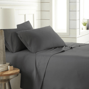 A neatly made bed with dark gray sheets and pillowcases is shown in a bright room with a side table holding a small plant and books.