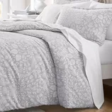A bedding set featuring a duvet cover and pillowcases with a subtle floral pattern in light grey tones, accented with white sheets underneath.