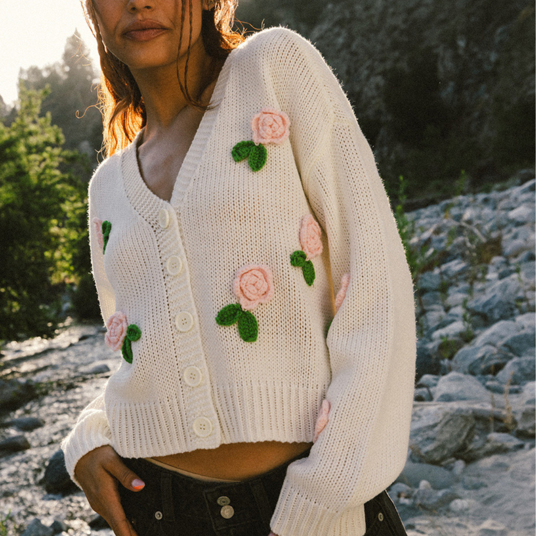 A woman wearing a white knitted cardigan with pink crocheted flowers and green leaves, featuring large white buttons. She stands outdoors with a natural, rocky background.