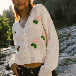 A woman wearing a white knitted cardigan with pink crocheted flowers and green leaves, featuring large white buttons. She stands outdoors with a natural, rocky background.
