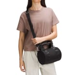 A person is wearing a brown t-shirt and beige pants, holding a small, black cylindrical bag with dual handles and a shoulder strap.