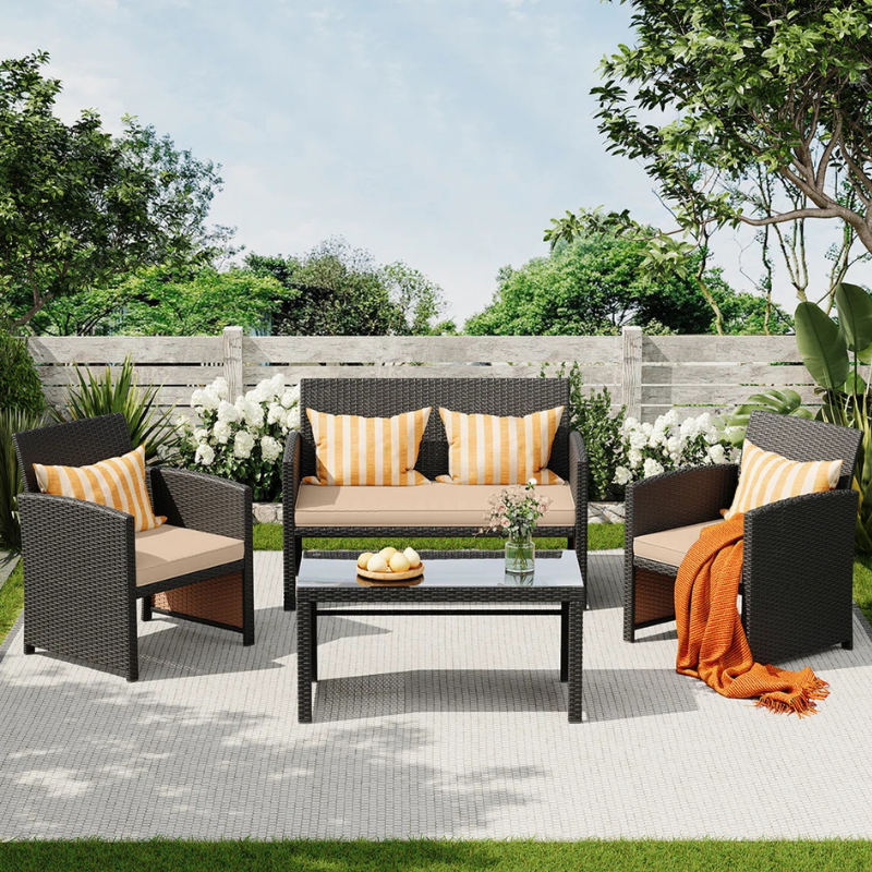 Outdoor patio set with a wicker loveseat, two chairs, and a glass-top coffee table. Beige cushions and orange-striped pillows add color, with a matching orange throw. Surrounded by greenery and a wooden fence.
