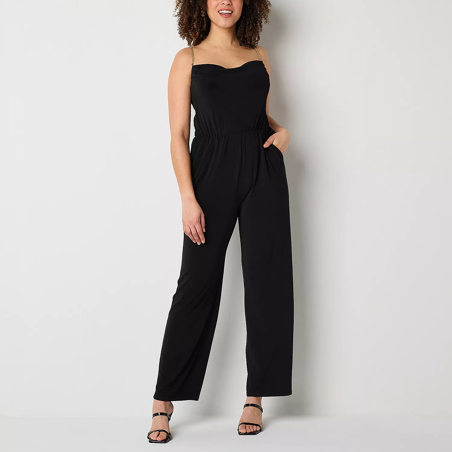 A woman models a sleeveless black jumpsuit with a straight neckline and high-waisted trousers, paired with black strappy sandals.