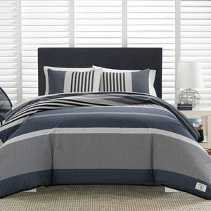 A Nautica Queen Comforter Set with a nautical color palette, featuring wide horizontal stripes in shades of navy, grey, and white, including two matching pillow shams.