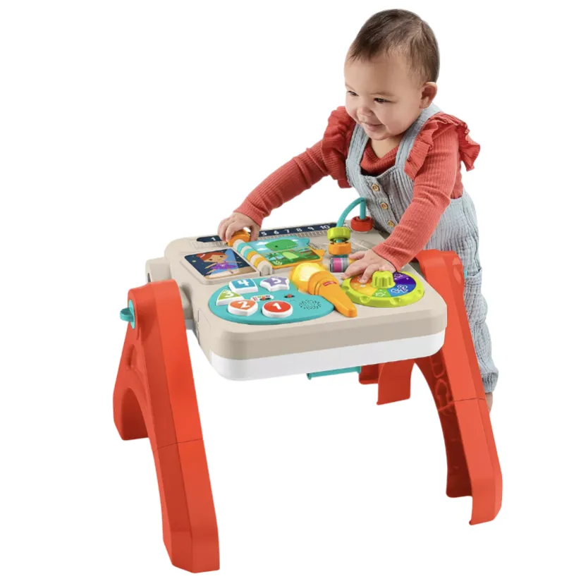 A child interacts with an activity table featuring various colorful buttons, switches, and dials designed for educational play. The table has red legs and a beige top with multicolored interactive elements.