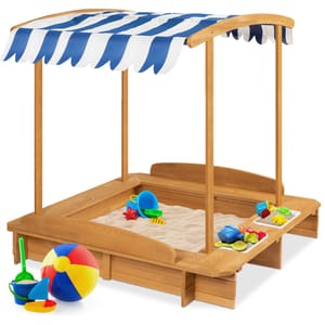 Wooden sandbox with a striped blue and white canopy and built-in benches, filled with sand and colorful toys including a beach ball.