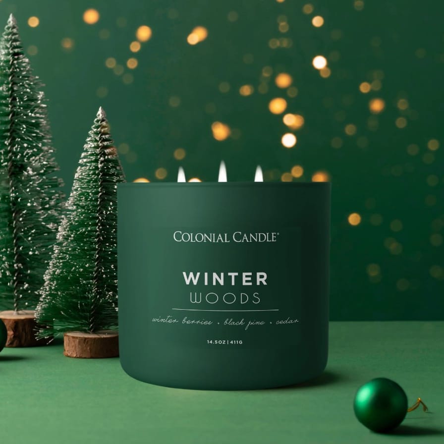 Colonial Candle \“Winter Woods\“ scented candle features aromas of winter berries, black pine, and cedar. The green candle is surrounded by small, snow-dusted pine trees and a green ornament, creating a festive atmosphere.
